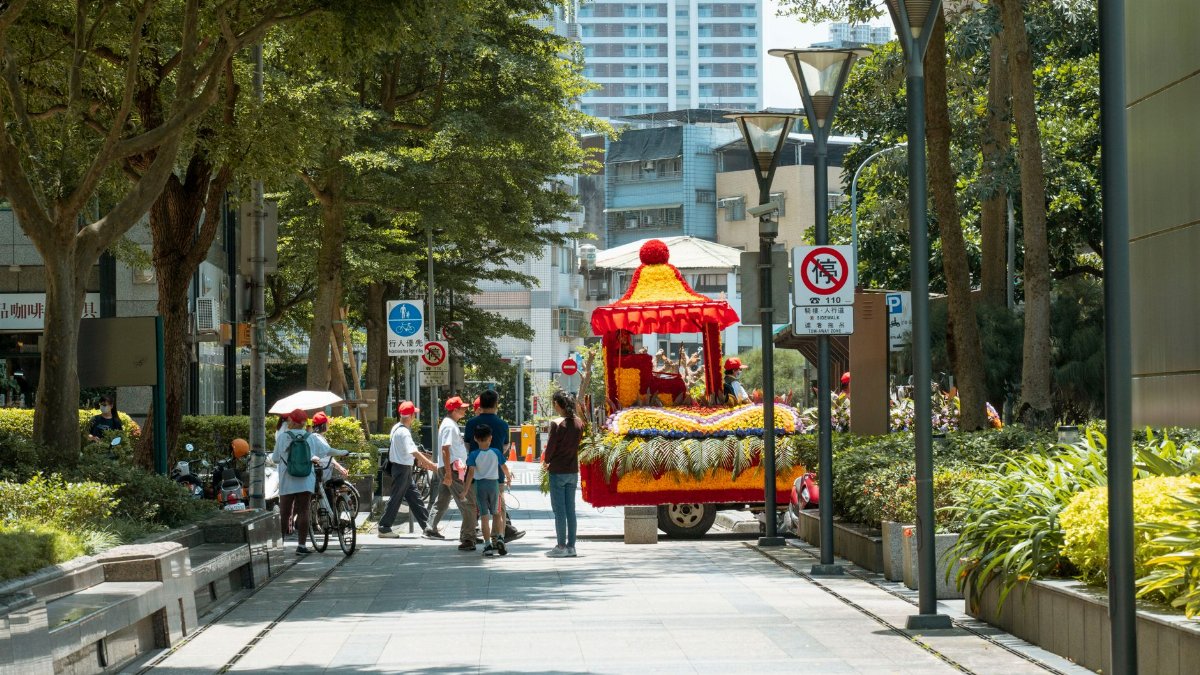 A vibrant parade float moves along a sunlit urban street lined with trees and pedestrians.
