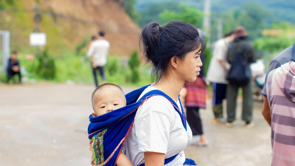 Young woman carrying baby on her back in a blue wrap outdoors, surrounded by nature.