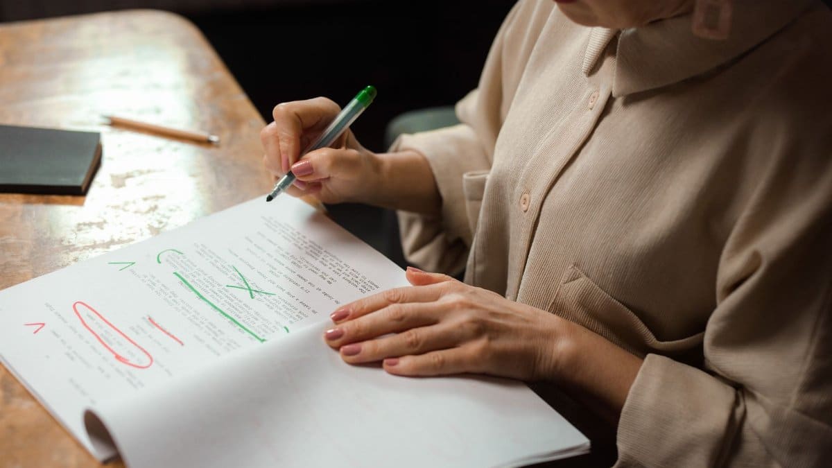 Close-up of a senior woman editing a manuscript at a desk with a green marker indoors.