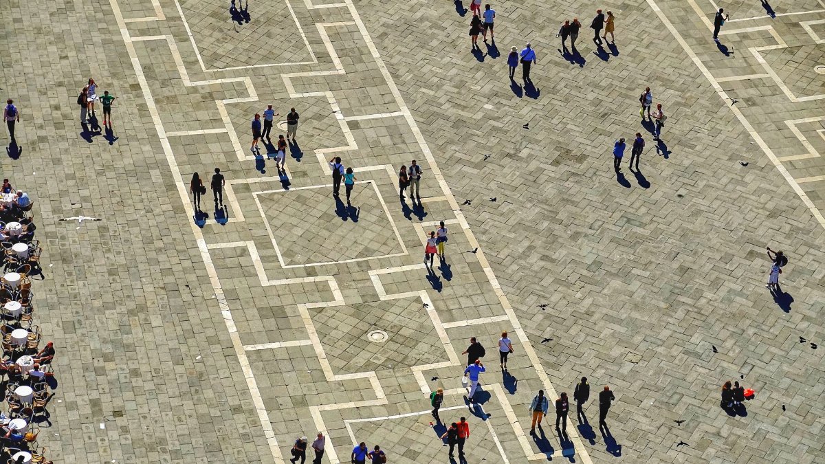 Aerial photo capturing people walking and socializing in St Mark's Square, Venice, with geometric patterns and long shadows.