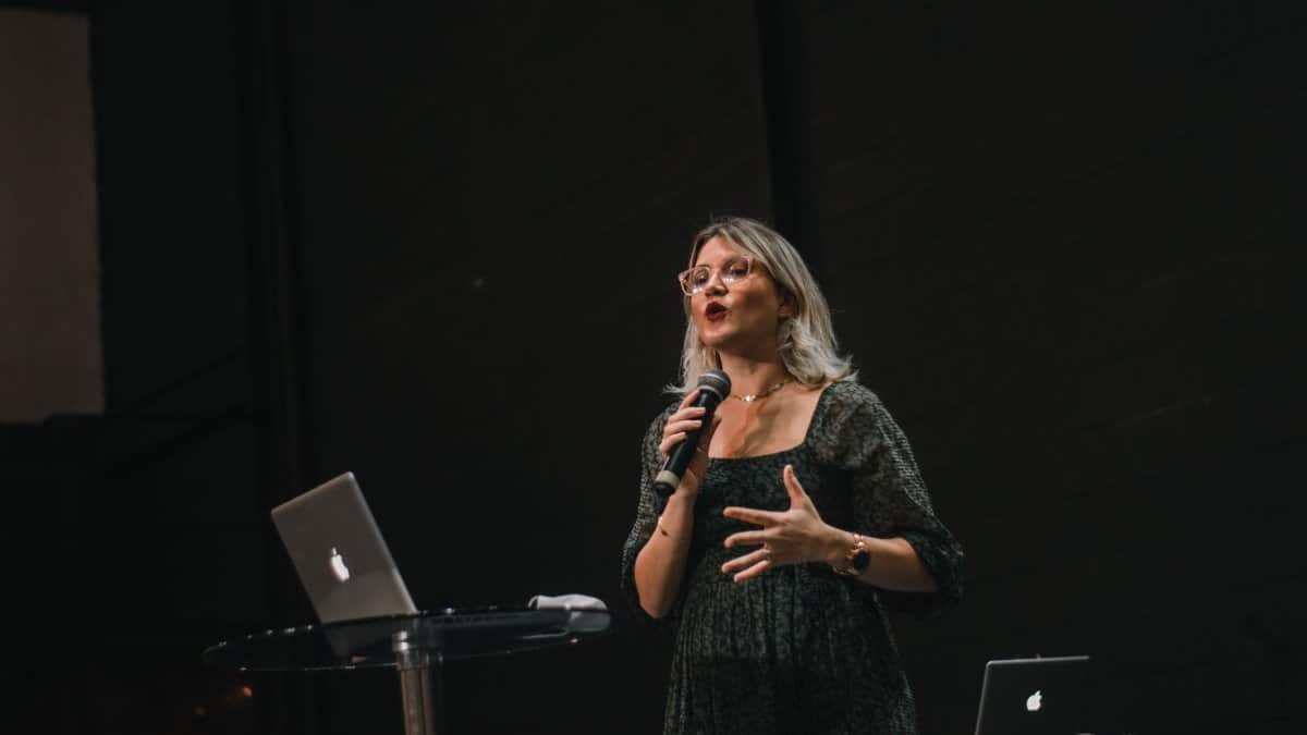 A confident woman delivers a speech on stage with a microphone and laptop.