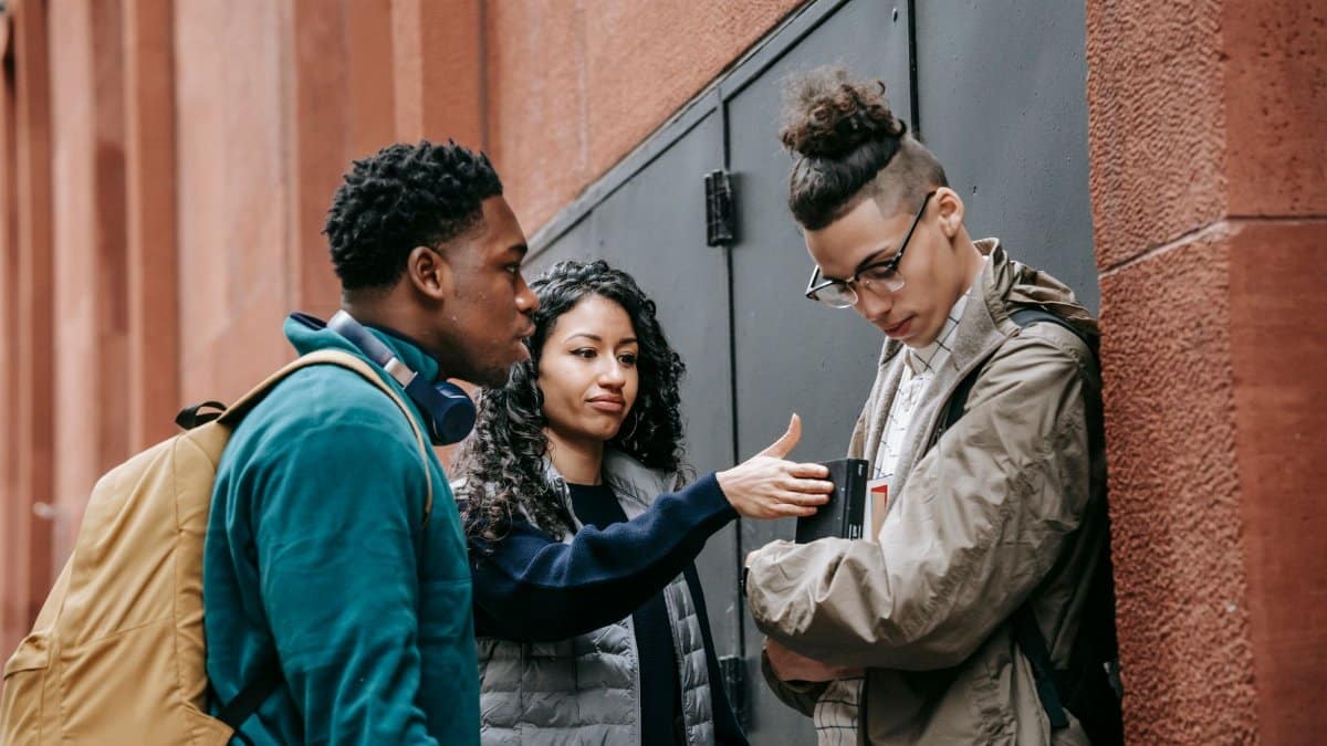 Multiracial young students in casual clothes with backpacks standing in city street while bullying male with books