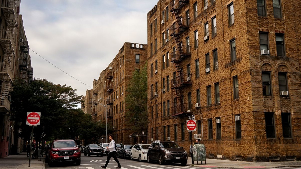 A city street with brick buildings, trees, cars, and a pedestrian crossing the street.