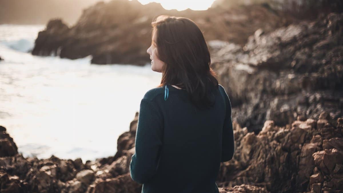 A woman stands thoughtfully by a rocky coastline at sunset, enjoying nature.