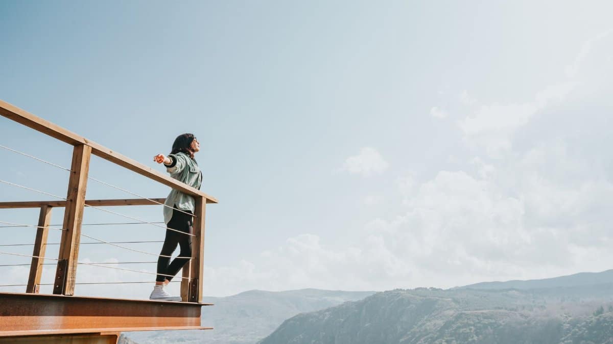 A woman relishes the adventurous mountain view from a scenic deck under a bright blue sky.