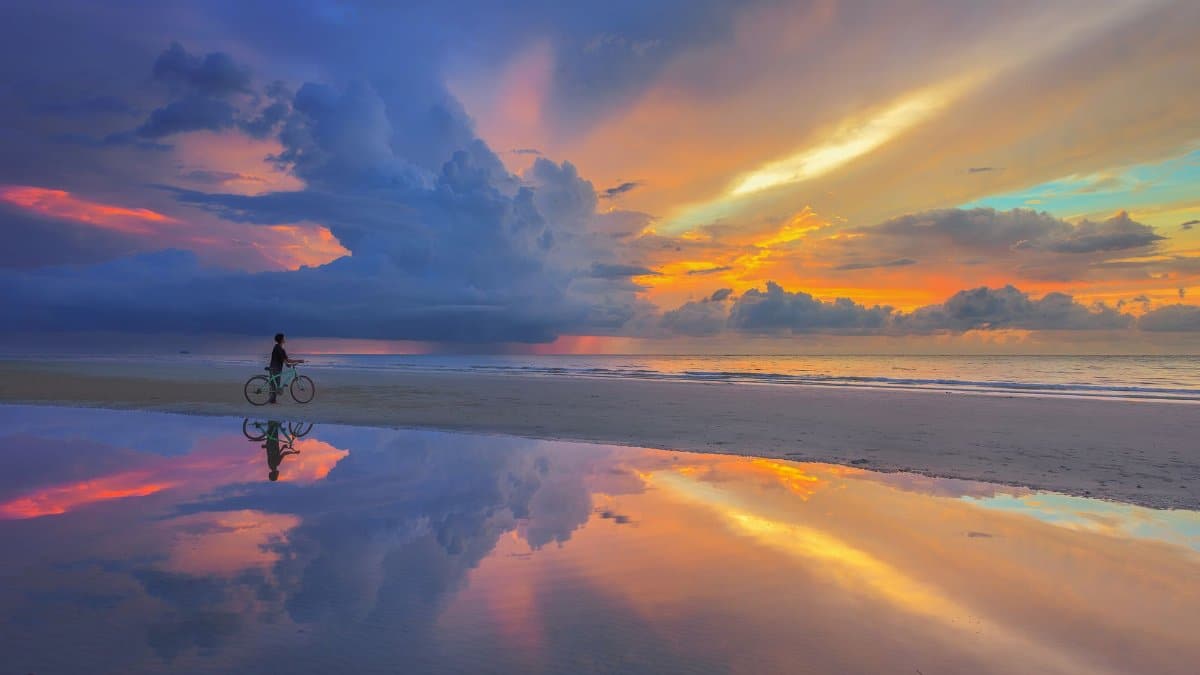 A person cycles along a beach during a stunning sunset with vibrant sky reflections.