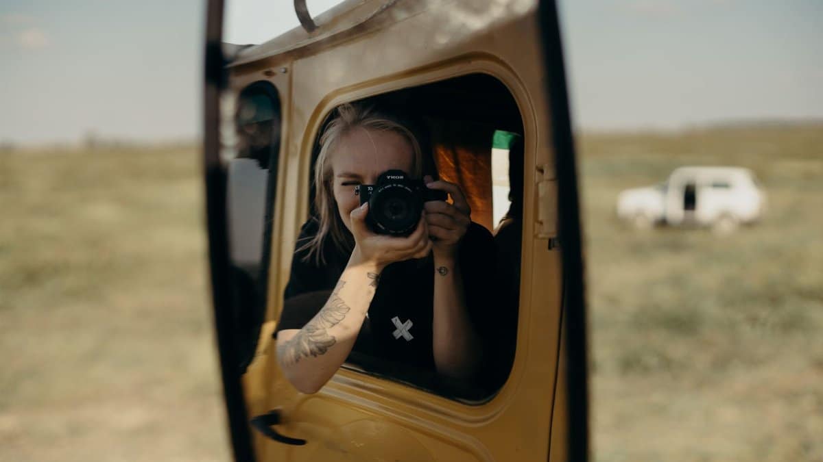 A woman with tattoos photographs her reflection in a vintage car mirror, embodying travel and nostalgia.