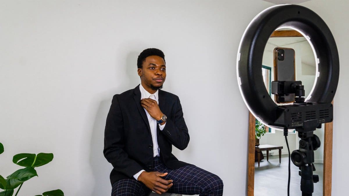 Young man adjusts outfit for a vlog in a modern setup with smartphone and ring light.