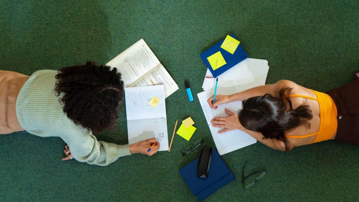 Top view of two women studying on a green carpet, with books and notes, embracing college education.