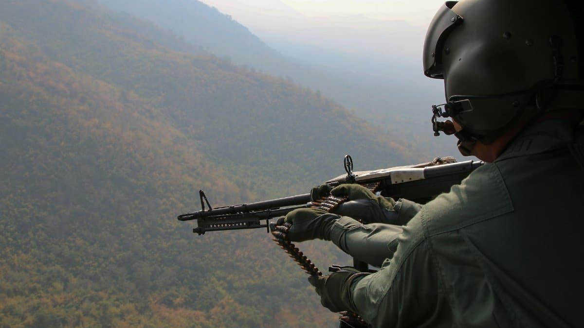 A soldier operates a machine gun from a helicopter above a vast landscape.
