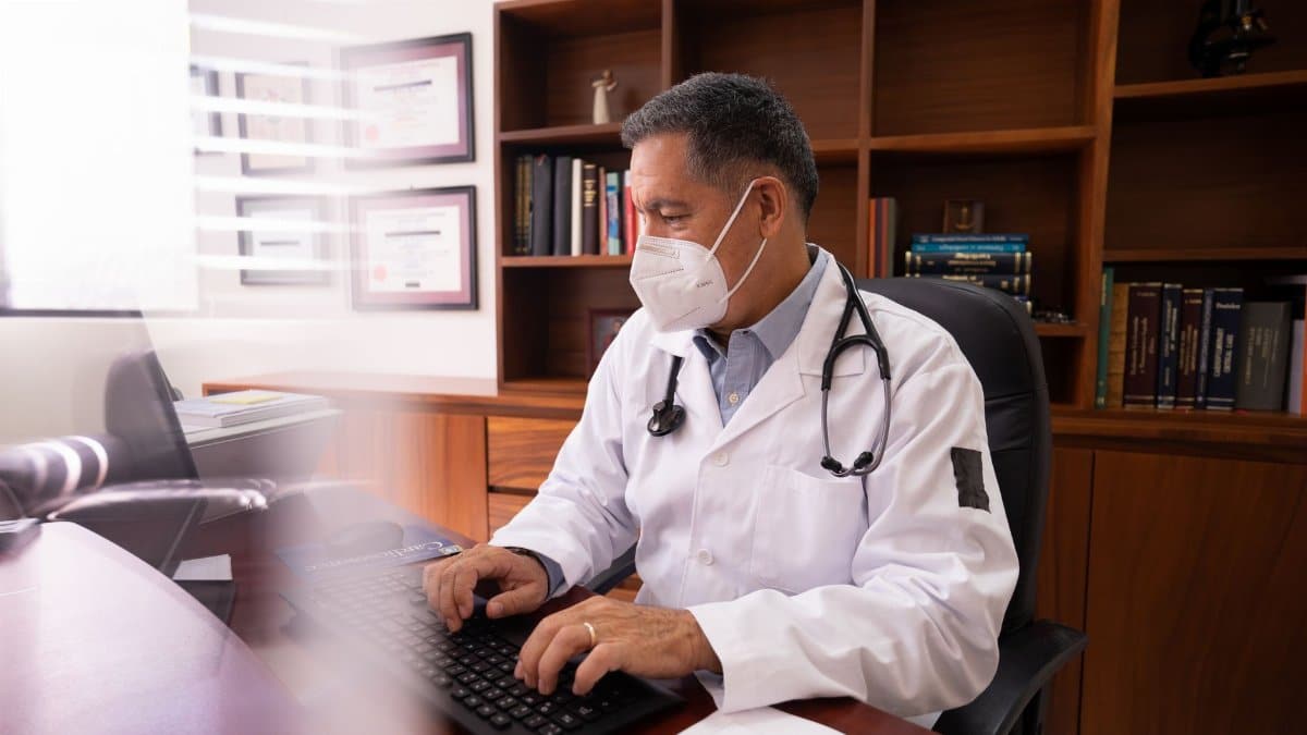 Medical professional in white coat with mask typing at desk in office setting.