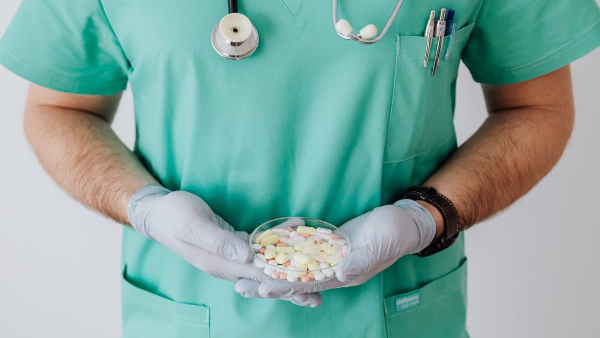 Unrecognizable male doctor wearing medical uniform and latex gloves holding Petri dish filled with assorted drugs while standing near white wall in hospital
