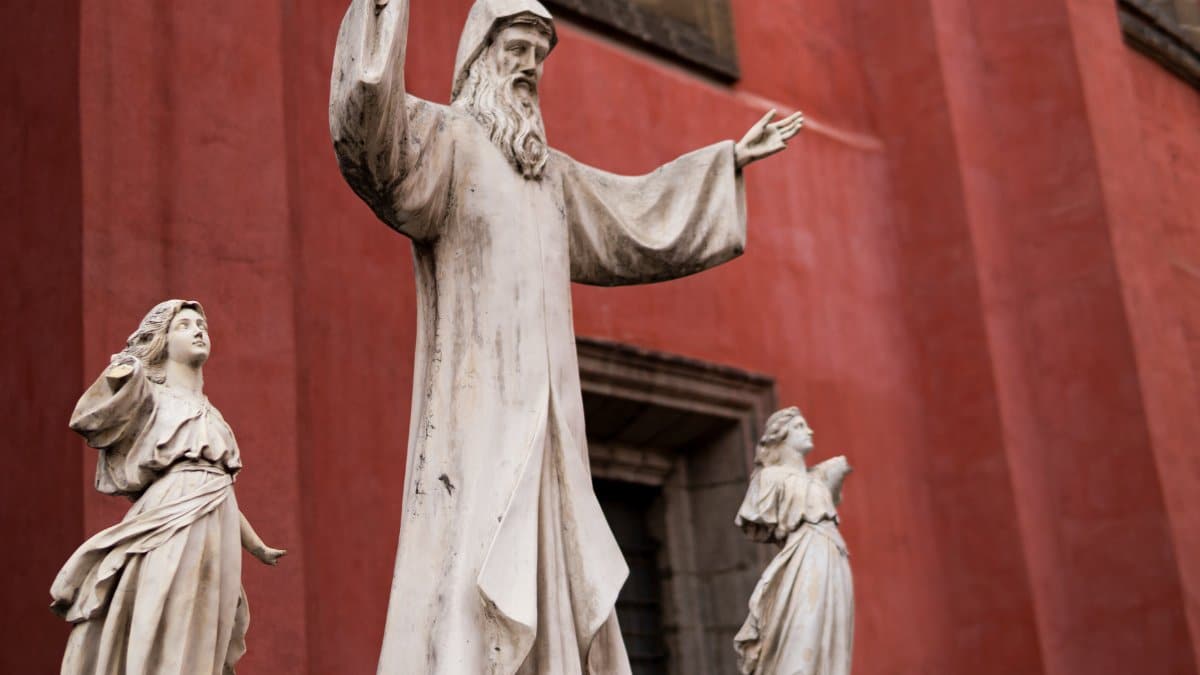 Marble religious sculptures of figures with raised arms on red wall backdrop.