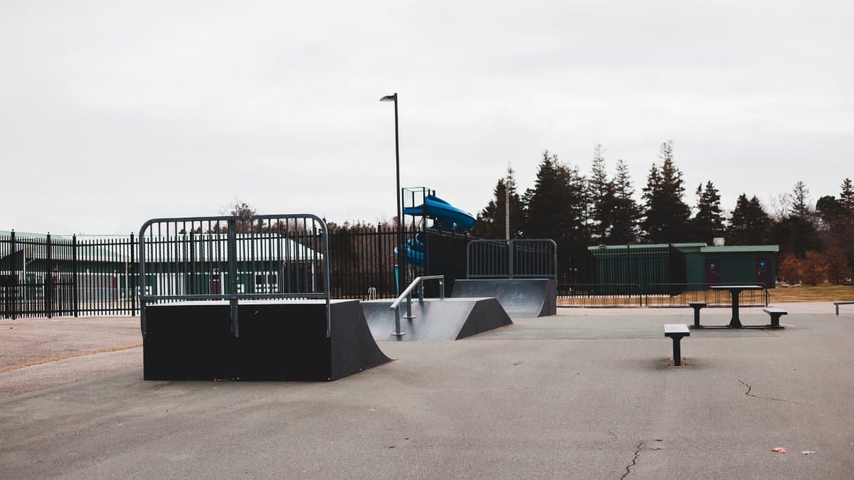 Empty ramps on asphalt paved street in skate park near benches and trees under cloudy gray sky in daytime
