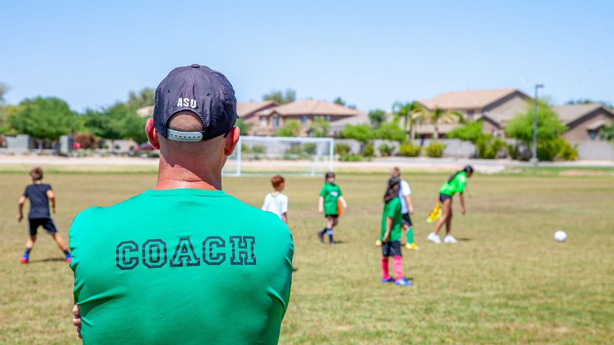 A soccer coach in a cap watches over children practicing on a sunny day at an outdoor field.