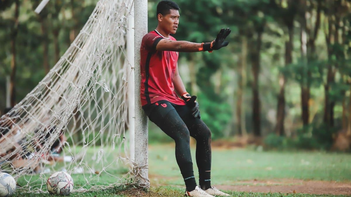 A soccer player taking a break by the goal post in a park, wearing a red jersey.