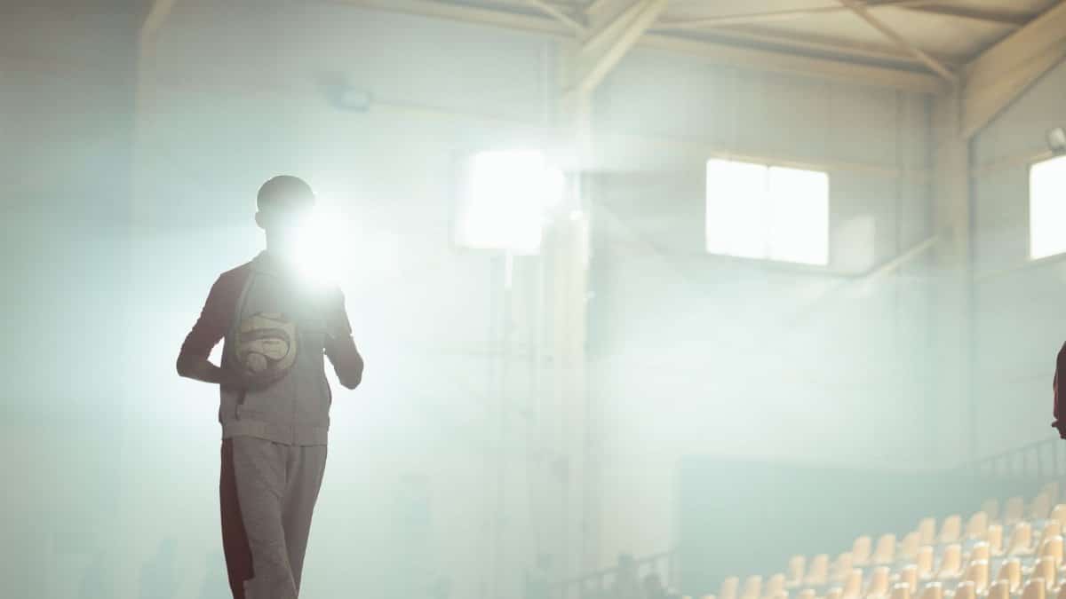 A silhouette of a basketball player holding a ball in a sunlit indoor sports arena.