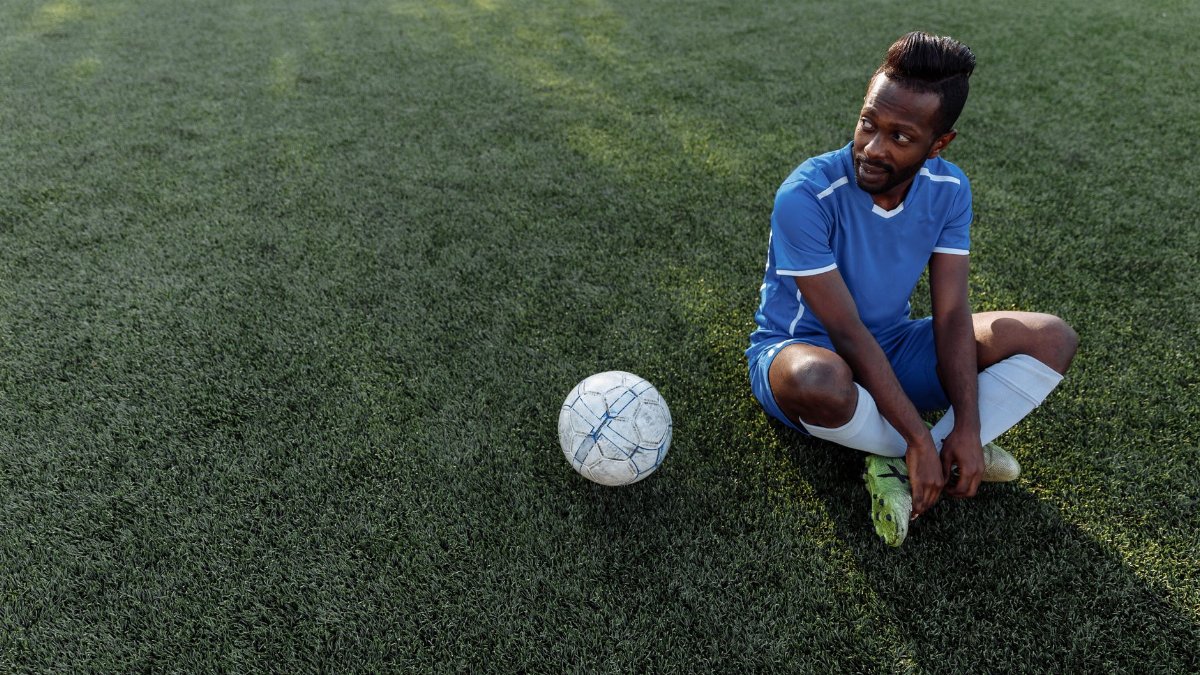 A soccer player in a blue jersey sits on the grass field during the day.
