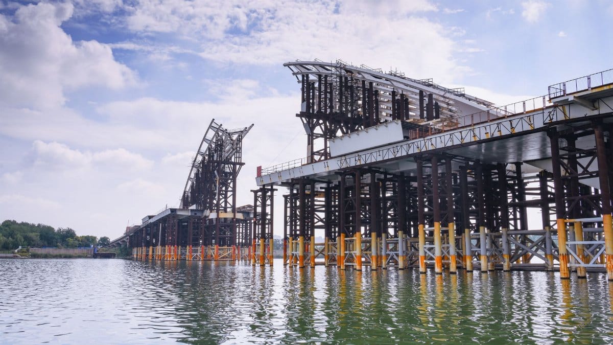 Steel framework of a large bridge under construction across a river on a sunny day.