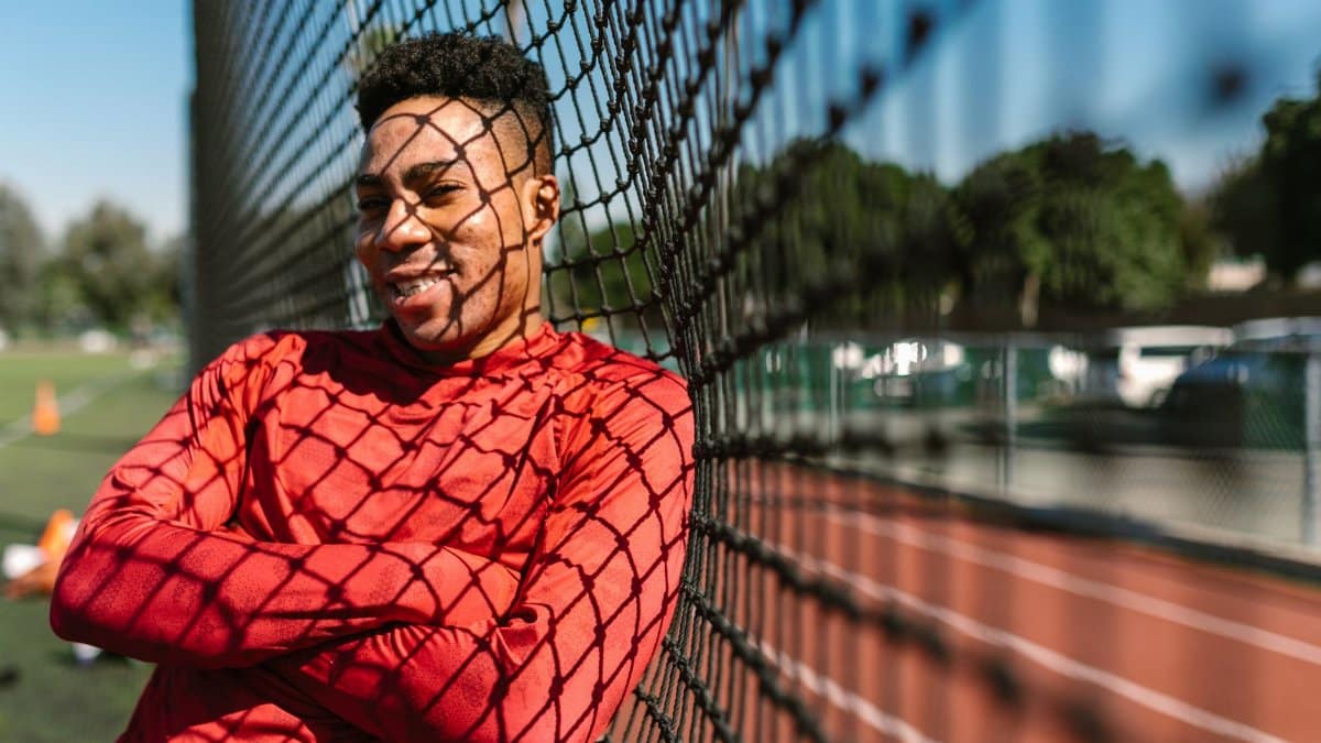 A cheerful athlete in a red shirt leaning on a sports field fence on a sunny day.