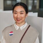 Asian woman smiling at polling station with 'Vote' badge on Voting Day.