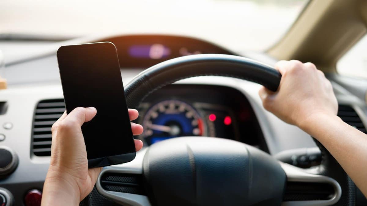 A driver uses a smartphone while steering a vehicle, showcasing distracted driving risks.