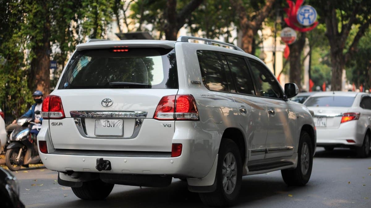 White SUV driving through an urban area with trees lining the street.