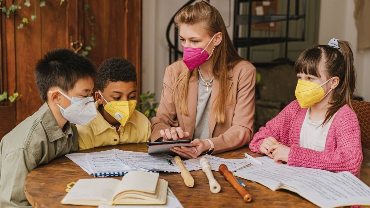 A group of diverse children learning music with a teacher indoors, using recorders and following safety measures.