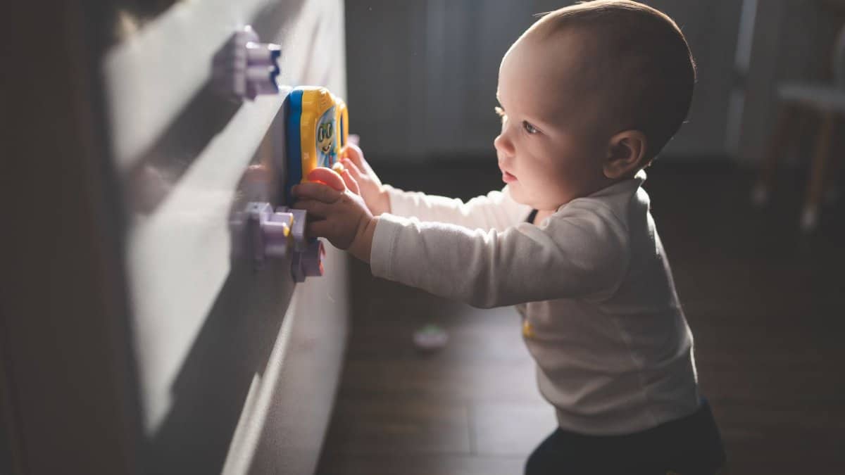 A cute baby exploring magnetic toys on a cupboard, illuminated by natural light indoors.