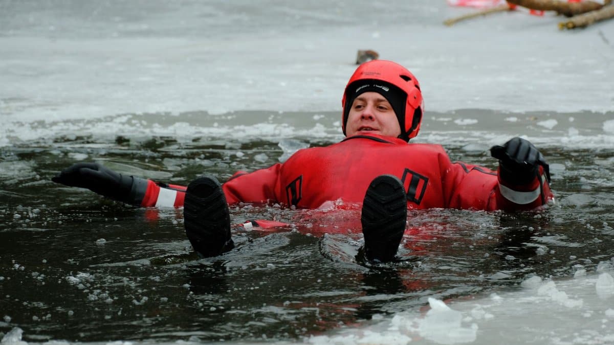 Adult male practicing ice rescue techniques in a red survival suit on frozen lake.