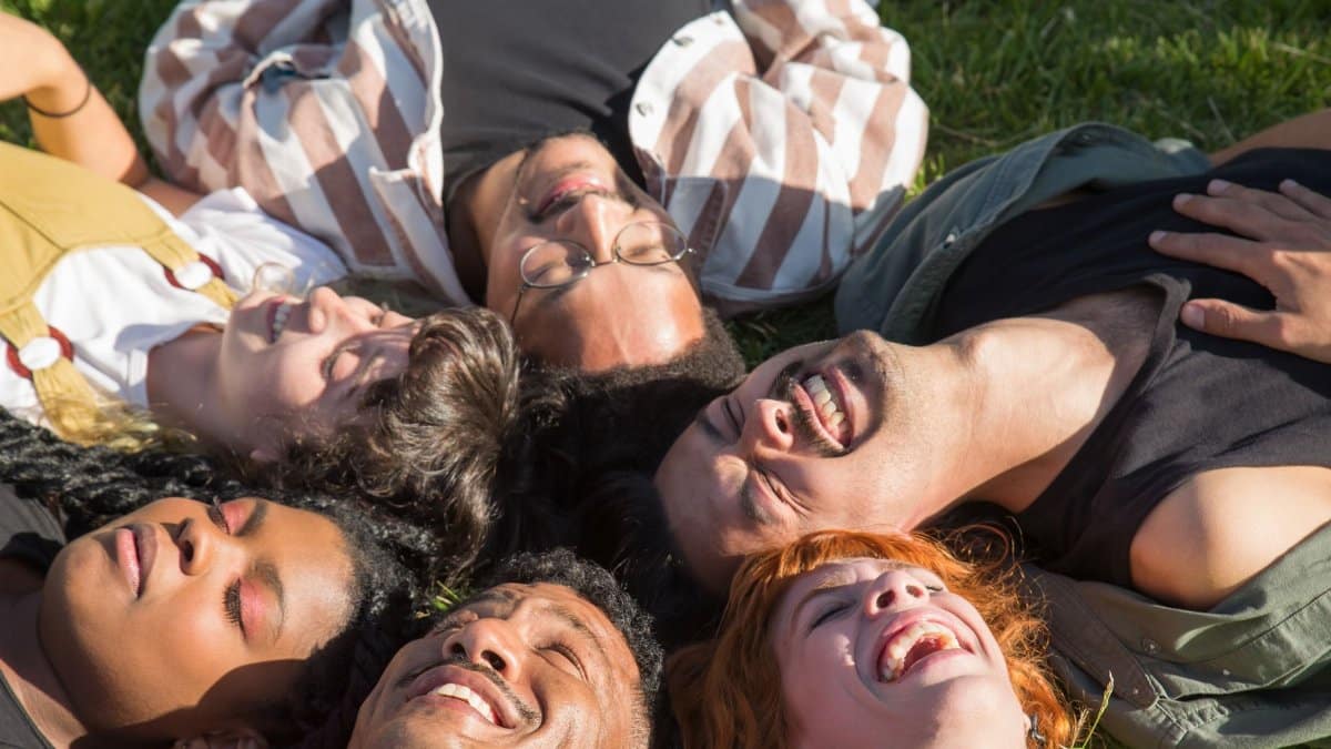A diverse group of friends lying in the grass, laughing under the sunny sky, showcasing joy and togetherness.