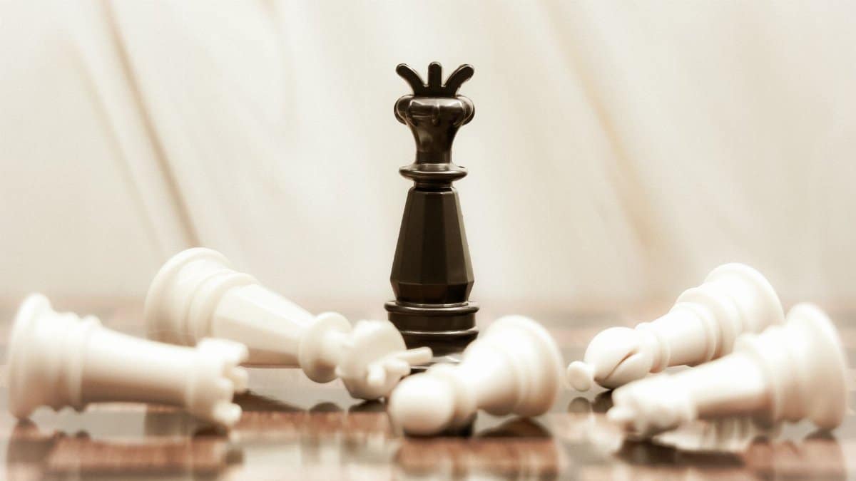 A black chess queen stands triumphant among toppled white pieces on a reflective surface.