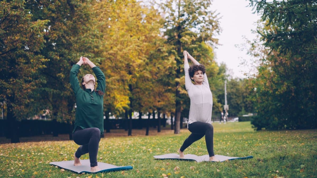 Two women practice yoga poses on mats in an autumn park environment.