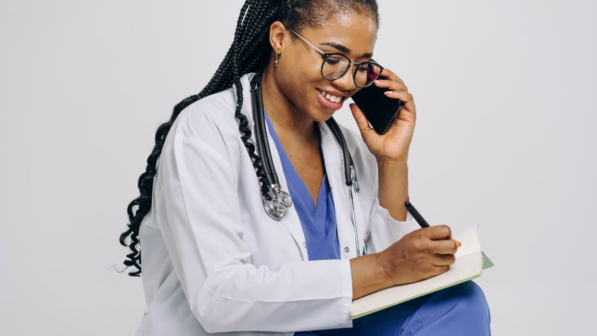 Female doctor smiling while talking on the phone and taking notes, wearing a stethoscope.