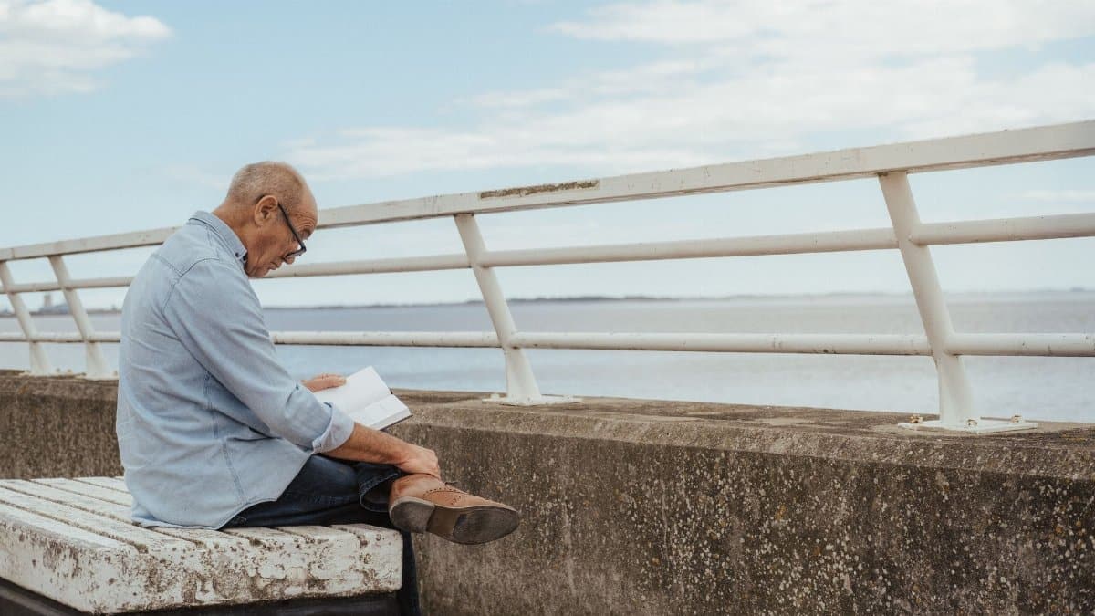 Side view of serious aged male wearing casual clothes and eyeglasses engaged in hobby reading book on embankment