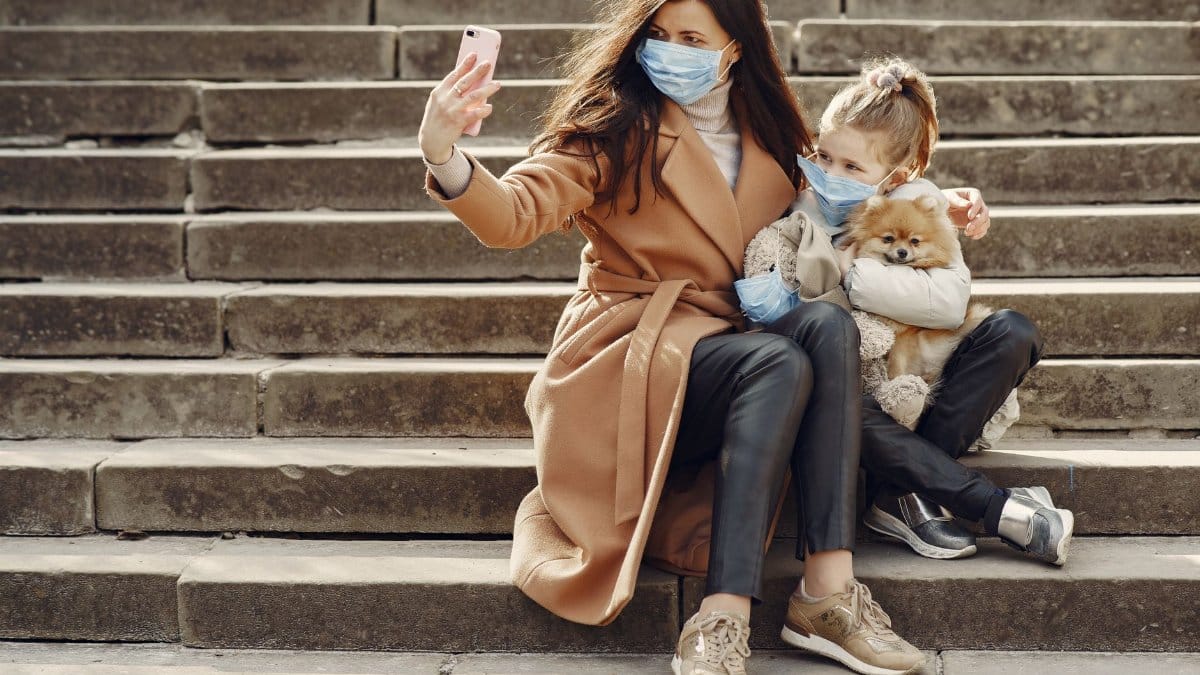 Full body of female and adorable little girl in casual clothes and medical masks sitting on steps with toy in mask and cute dog taking photo on smartphone