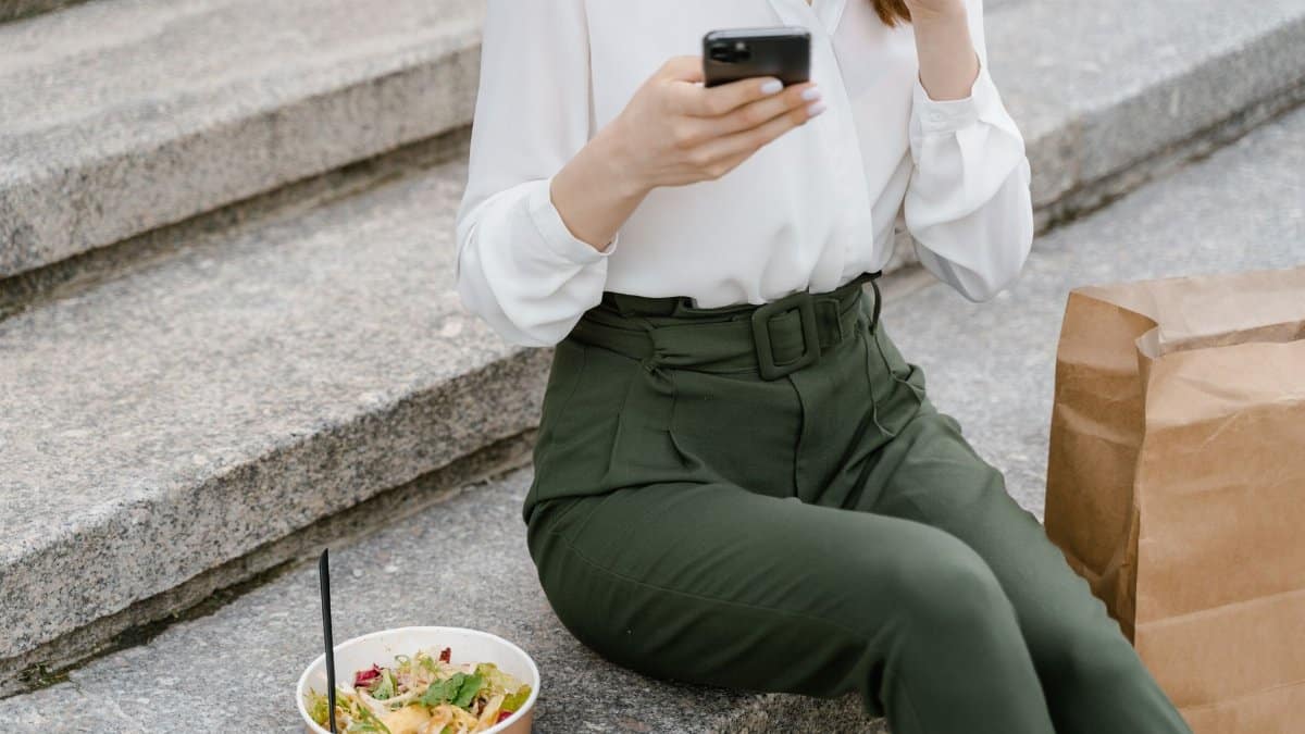 A woman sits on concrete steps with a salad and paper bag, using a smartphone.
