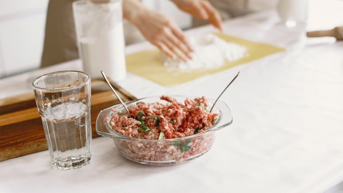 Raw ground meat in a glass bowl with ingredients being prepared nearby.