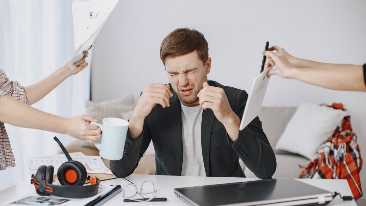 Adult man stressing out at home office desk with multiple demands surrounding him.