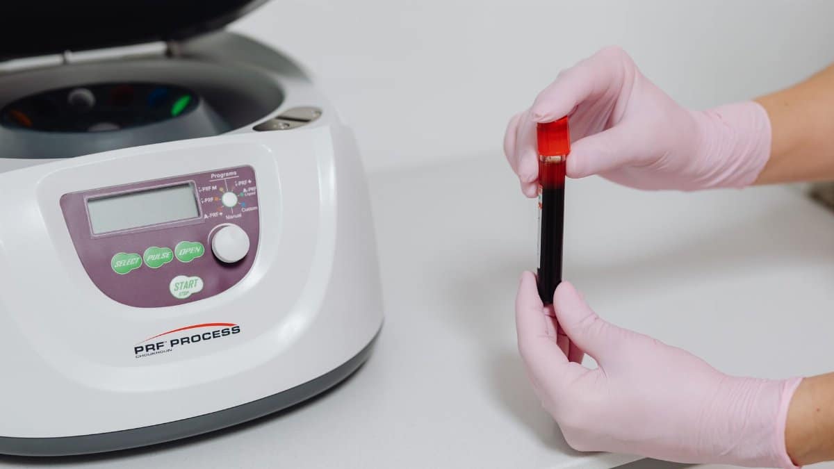 Close-up of hands in gloves holding a blood sample beside a centrifuge.