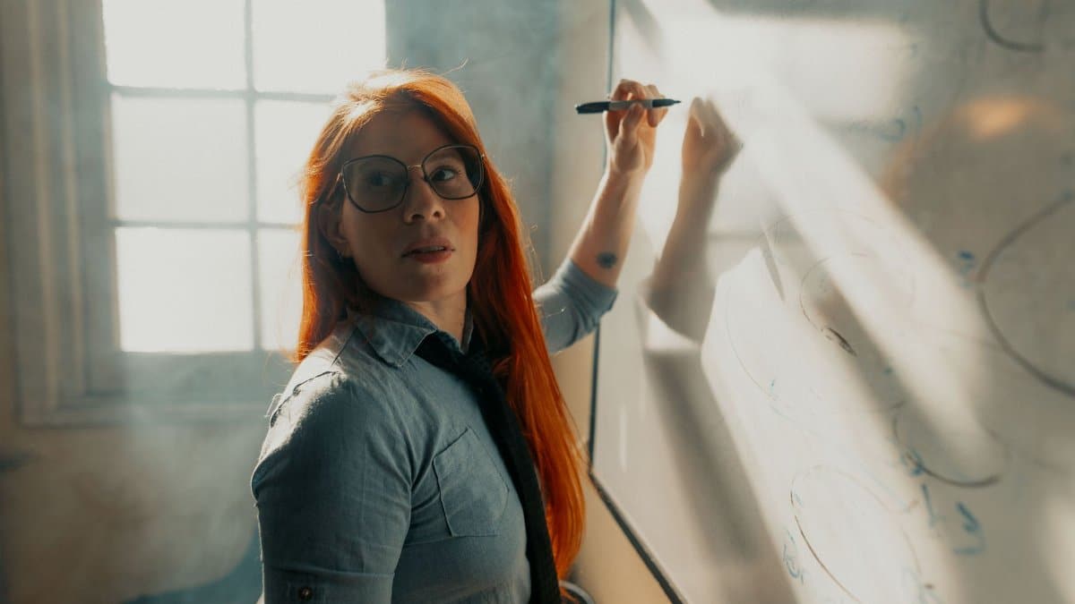 Red-haired woman writing on whiteboard in a sunlit room, exuding confidence and focus.