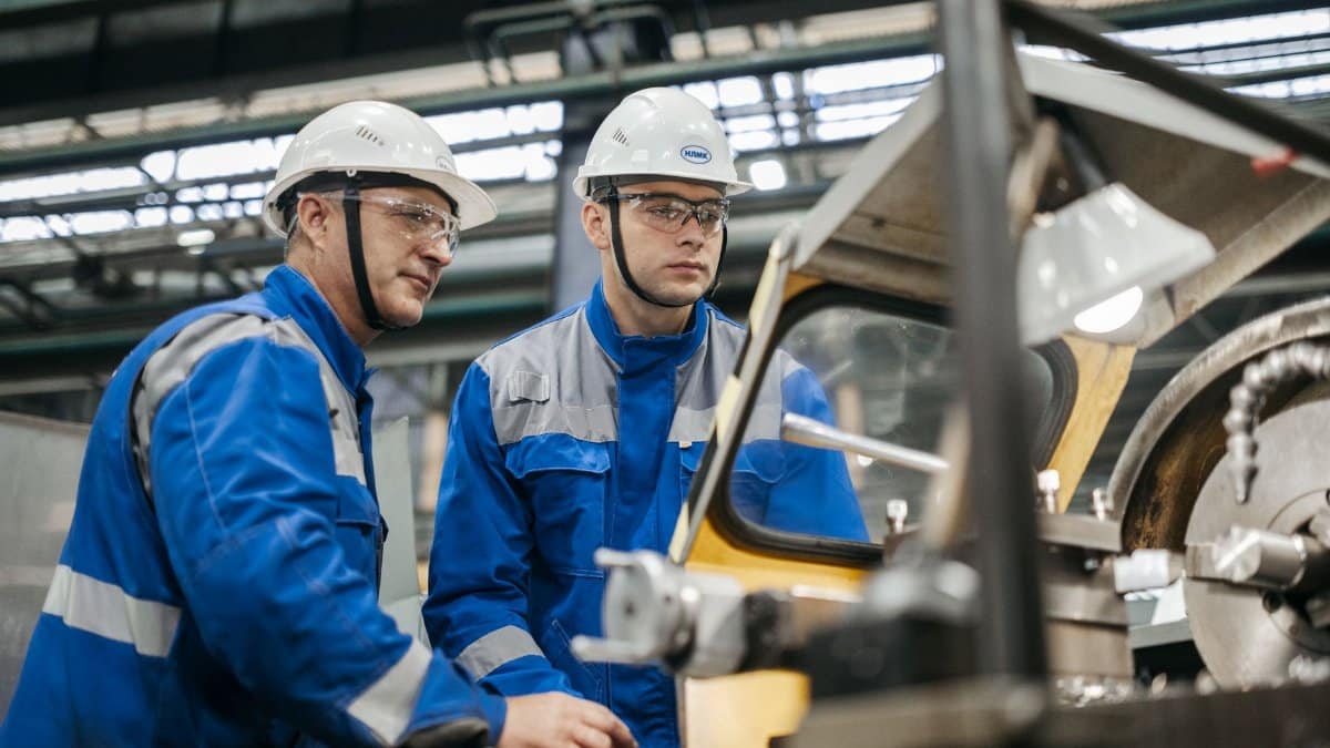 Two industrial workers in protective gear operate machinery in a factory setting.