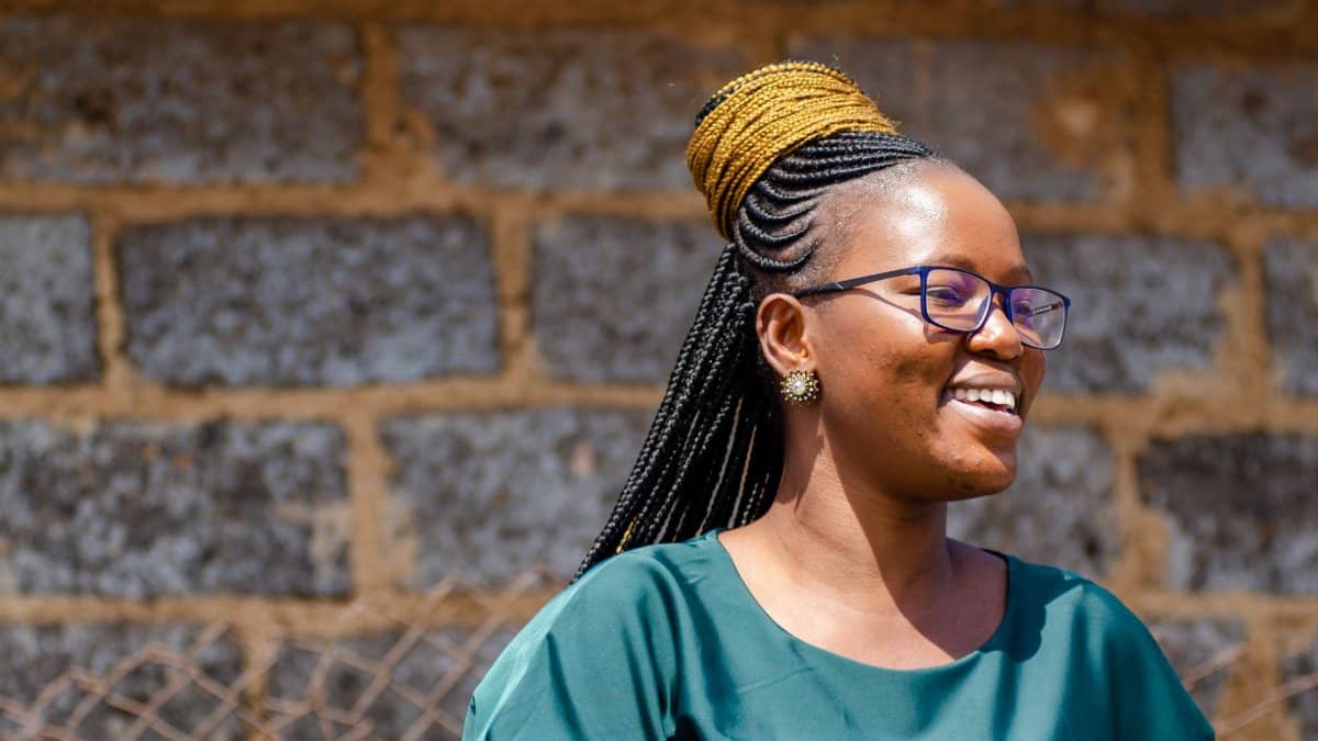 A cheerful woman with braided hair and glasses smiles outdoors against a stone wall.