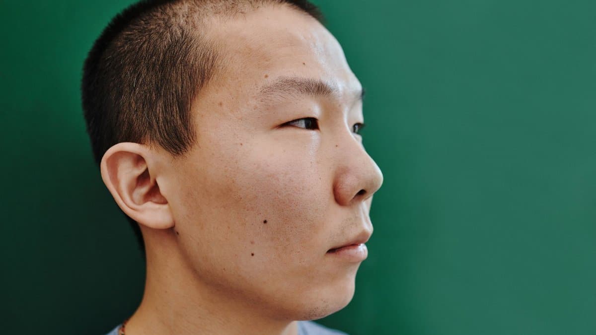 Close-up profile of an East Asian man against a green background, showcasing short hair.