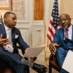 Two African American men in formal attire discussing documents in an office setting with flags in the background.