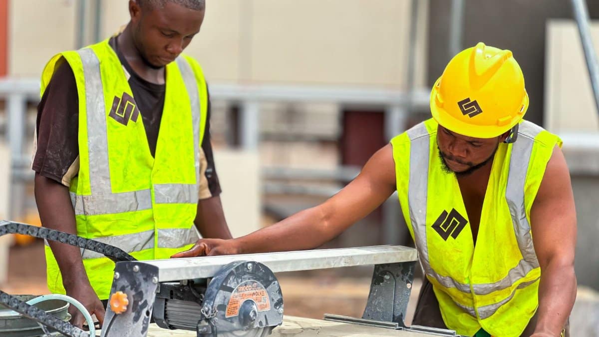 Two construction workers using a tile saw on site, wearing safety gear and high-visibility vests.