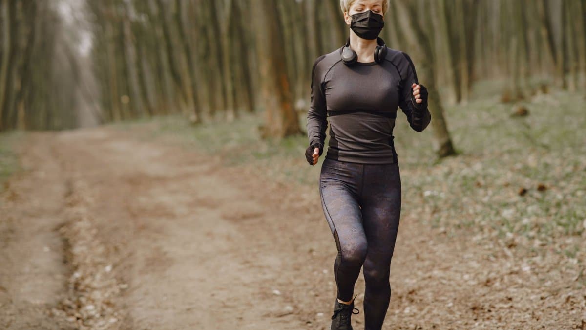 Full body sportswoman in activewear and medical mask with headphones on neck running through autumn forest with closed eyes during workout