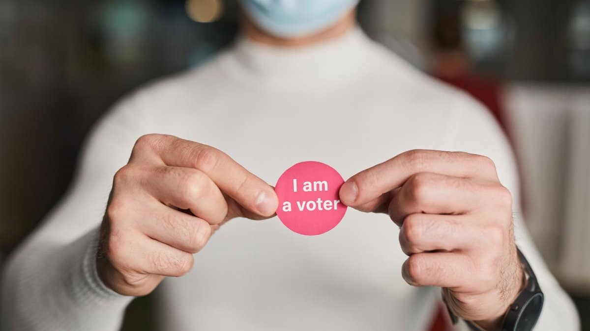 Person in a mask holding a voter sticker, symbolizing civic engagement.
