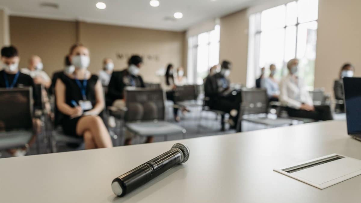 A business conference room setting with masked participants and a microphone in the foreground.