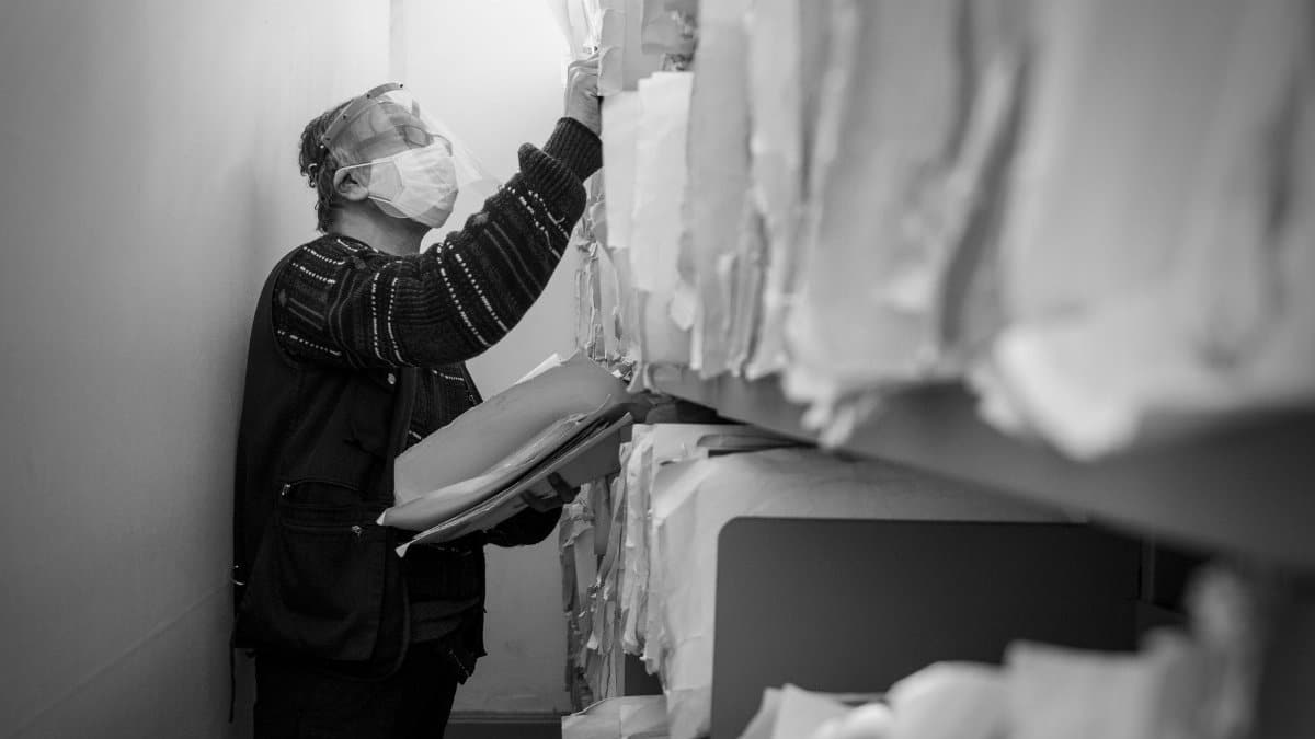 Black and white image of a masked elderly man organizing records in an office aisle.